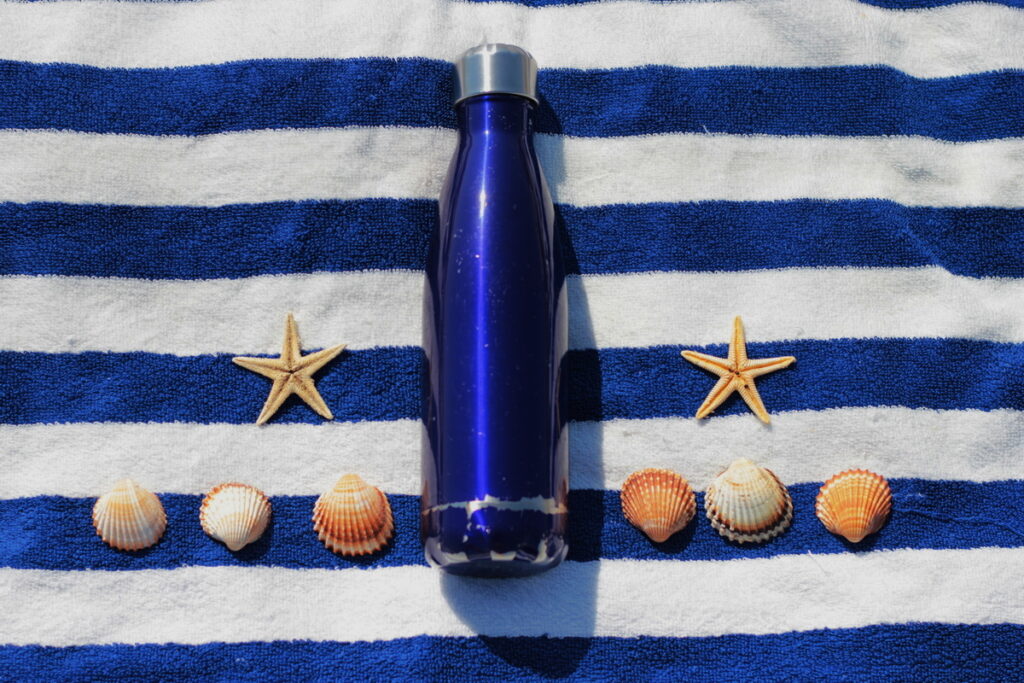 Blaue Trinkflasche auf gestreiftem Tuch, flankiert von Muscheln und Seesternen in einer ruhigen Strandkomposition.