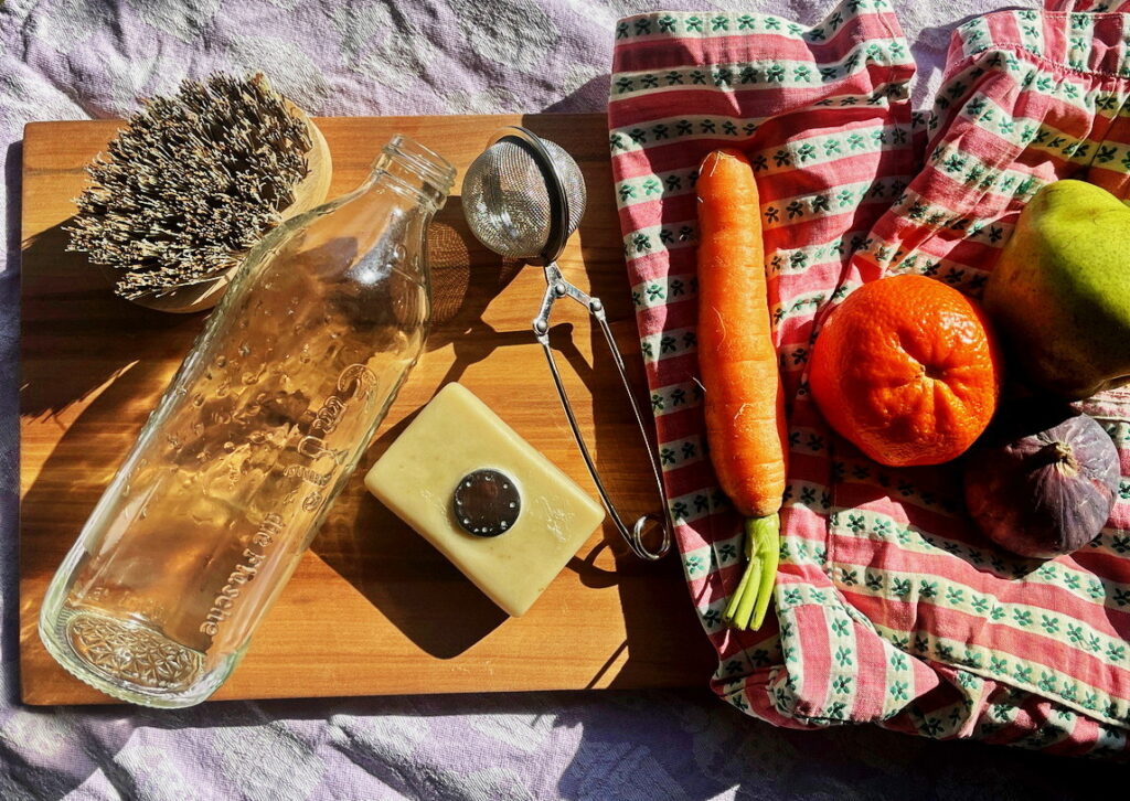 Flatlay mit Glasflasche, Seifenstück mit Magnethalter, Holzbürste, Teesieb, Karotte, Mandarine, Birne und Feige auf einem Holzbrett und Stoff